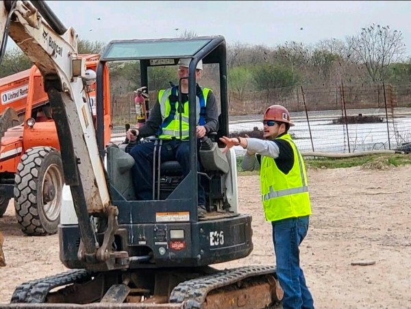 Instructor giving hands-on excavator operation guidance at an excavation site